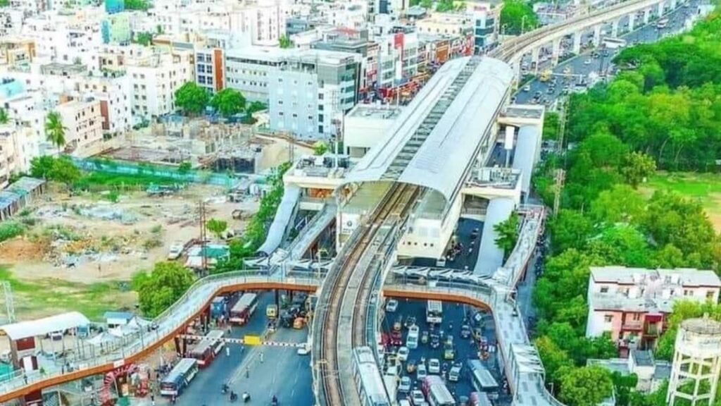 "Aerial view of elevated metro rail station with curved platform roof, surrounded by urban buildings, green trees, and busy road traffic below"