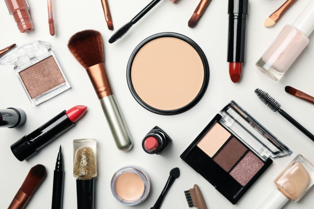 "Flat lay of makeup essentials including lipstick, eyeshadow palette, brushes, mascara, and nail polish on white background"